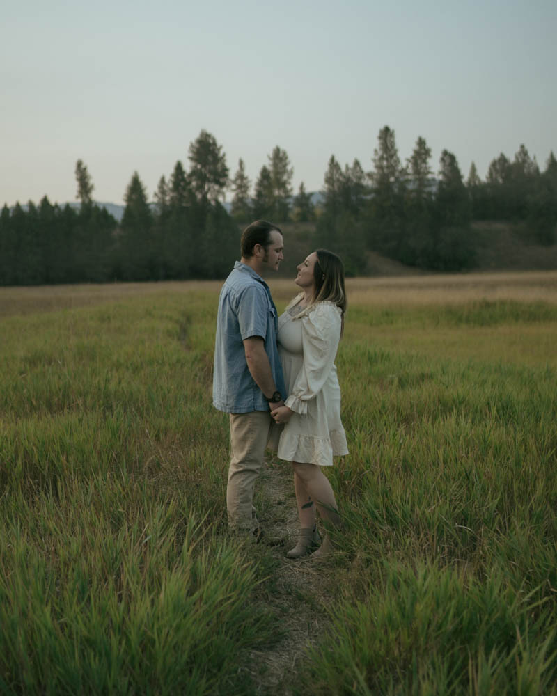 couple standing on a trail surrounded by tall grass as they hold hands and look at each other.