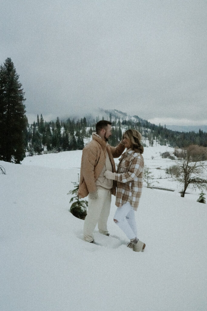 couple standing on a snow covered hill side looking at each other with the mountains in the bcakground.  