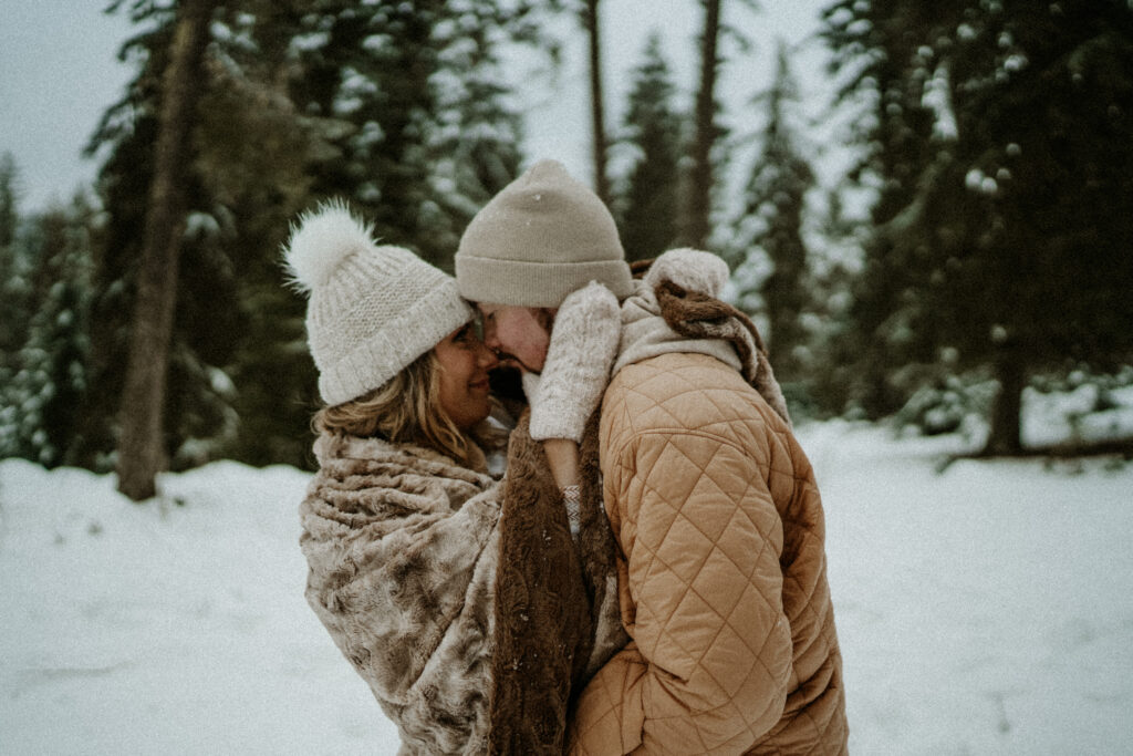 couple cuddling close in their winter hats and coats outdoors in the snow covered woods of Coeur d' Alene, Idaho.  