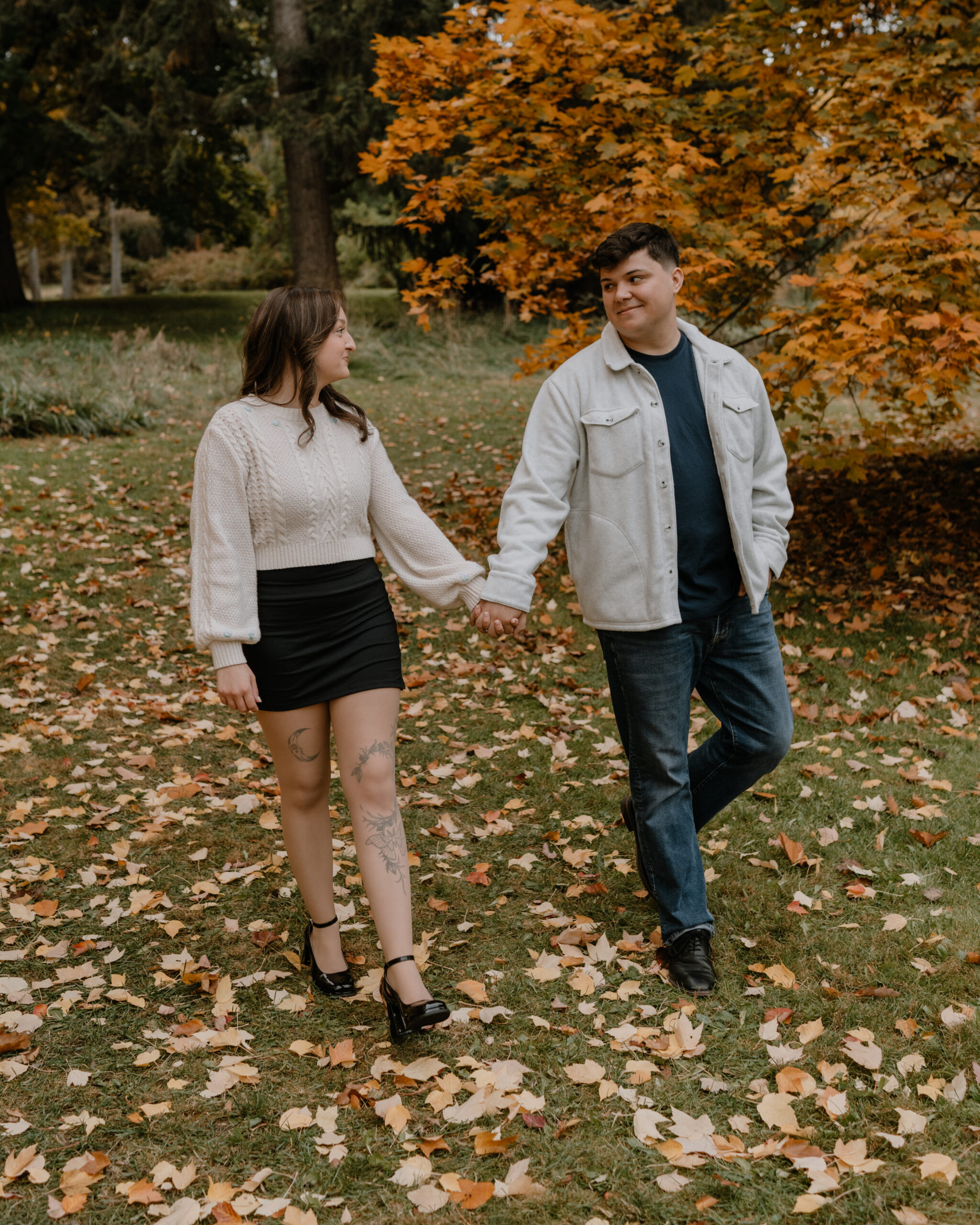 A couple walks hand in hand through a park during autumn, surrounded by fallen leaves and vibrant orange foliage. The woman wears a cream knit sweater and a black skirt, while the man wears a light-colored jacket over a dark shirt and jeans. They gaze at each other warmly, sharing a moment in the natural setting.