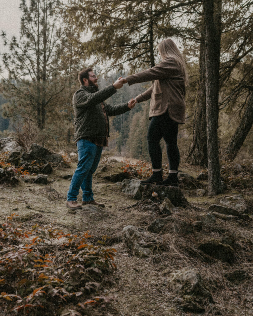 Idaho Winter Couples Session where the husband is holding his wife's hands as she steps off a tree stump.