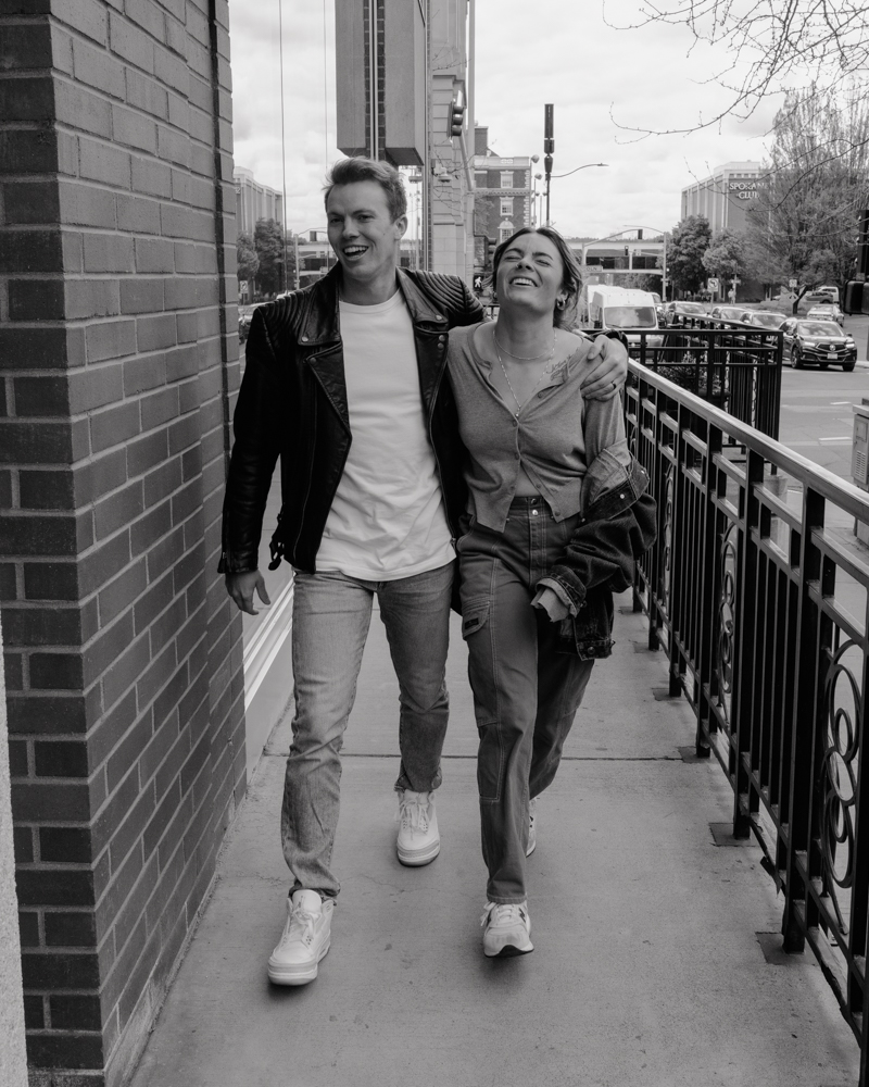 a couple walking arm in arm through downtown Spokane, laughing together. The man wears a leather jacket over a white shirt and jeans, while the woman wears a casual cardigan and cargo pants, holding a denim jacket. The urban background features city buildings, traffic, and a railing along the sidewalk, capturing a candid and joyful moment.