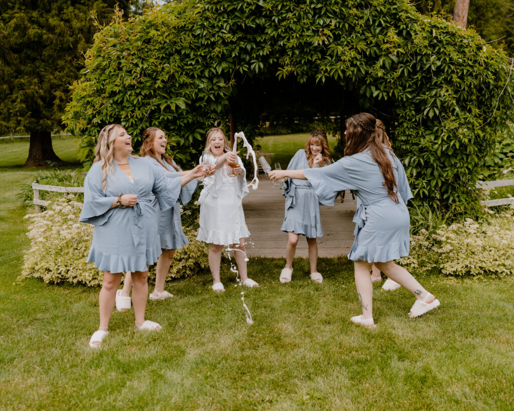 bride with her bridesmaids standing in front of an ivy covered bridge in their dusty blue robes with ruffles as the bride pops the champagne to toast to her wedding day and the bridesmaids are full of excitement.  