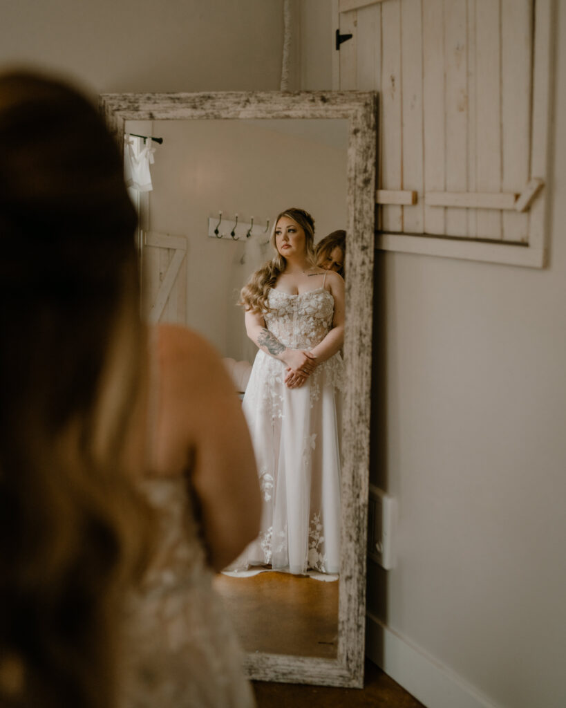 a bride's reflection in a rustic, weathered mirror as she prepares for her wedding. She stands with her hands gently clasped, gazing thoughtfully at her reflection. Her gown features intricate floral lace detailing, and her long, wavy hair cascades over one shoulder. A bridesmaid or helper is partially visible behind her, adjusting her dress. The setting is a cozy, farmhouse-style room with wooden accents, a sliding barn door, and soft natural light filtering in.