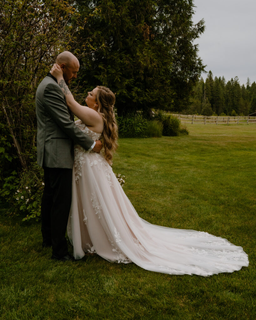 bride and groom standing and embracing near greenery with a rustic wooden fence in the background.  They are staring into each others eyes as the bride outstretches her arm to touch the grooms face. 