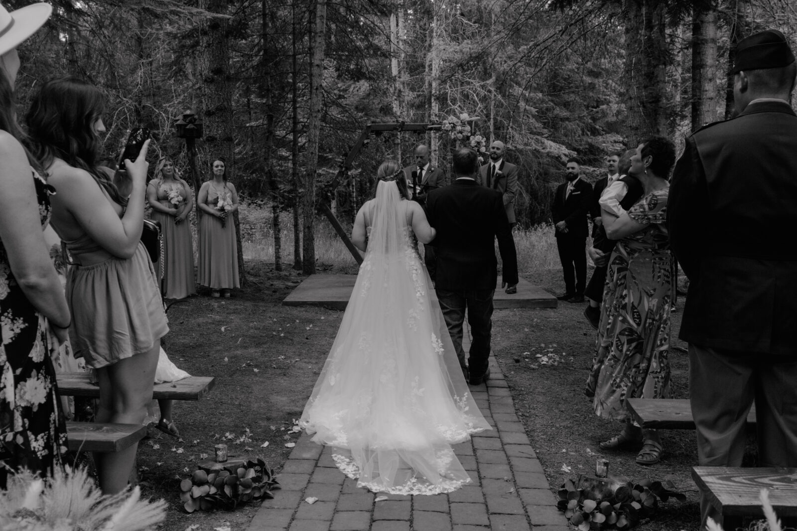 bride walking down the aisle with her escort at an intimate outdoor wedding ceremony in a forest setting. She wears a flowing gown with delicate lace details and a long veil trailing behind her. Guests stand on either side, some capturing the moment on their phones, while bridesmaids in matching dresses wait near the altar. The groom and officiant stand beneath a floral-adorned geometric arch, surrounded by tall trees, creating a serene and romantic atmosphere.