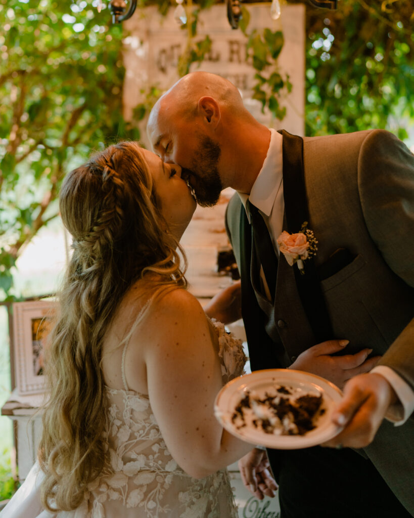 bride and groom standing in front of their cake as they kiss.  The groom holding a plate full of cake in his hand. 