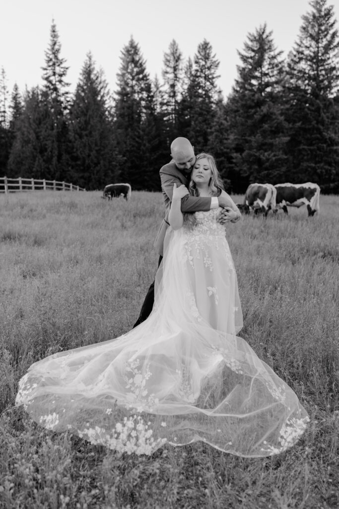 newlywed couple embracing in a pastoral field. The groom, dressed in a suit, stands behind the bride, wrapping his arms around her shoulders as he gently leans his head toward her. The bride, wearing a flowing gown with delicate floral appliqués, closes her eyes with a serene expression. Her long veil cascades over the grass, creating an ethereal effect. In the background, cows graze in the open field, with a wooden fence and tall evergreen trees framing the scene.